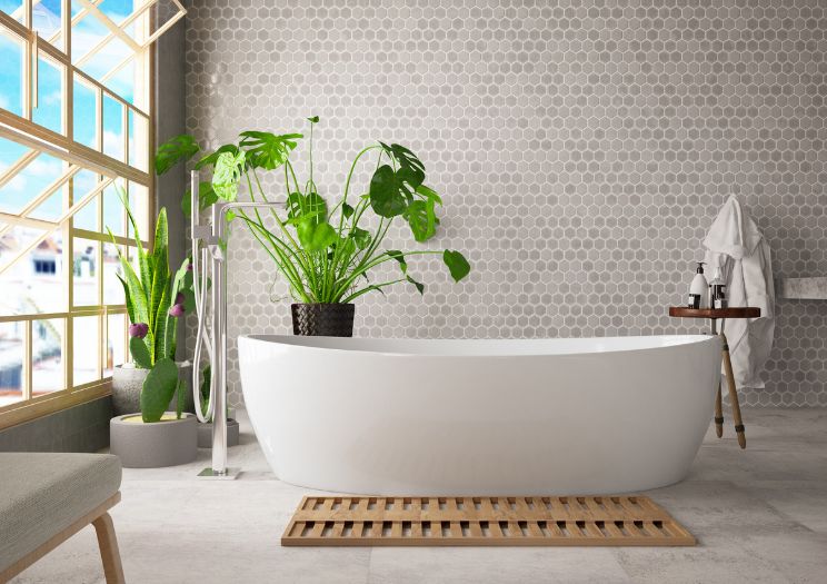 A grey tiled bathroom with a large green plant and porcelain bathtub is pictured in overlooking a window with blue sky.