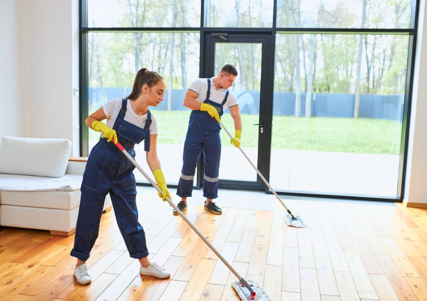 Two cleaners in navy overalls and yellow latex gloves hold mops as they push them across the hardwoor floor of a rental unit for a cleaning service.